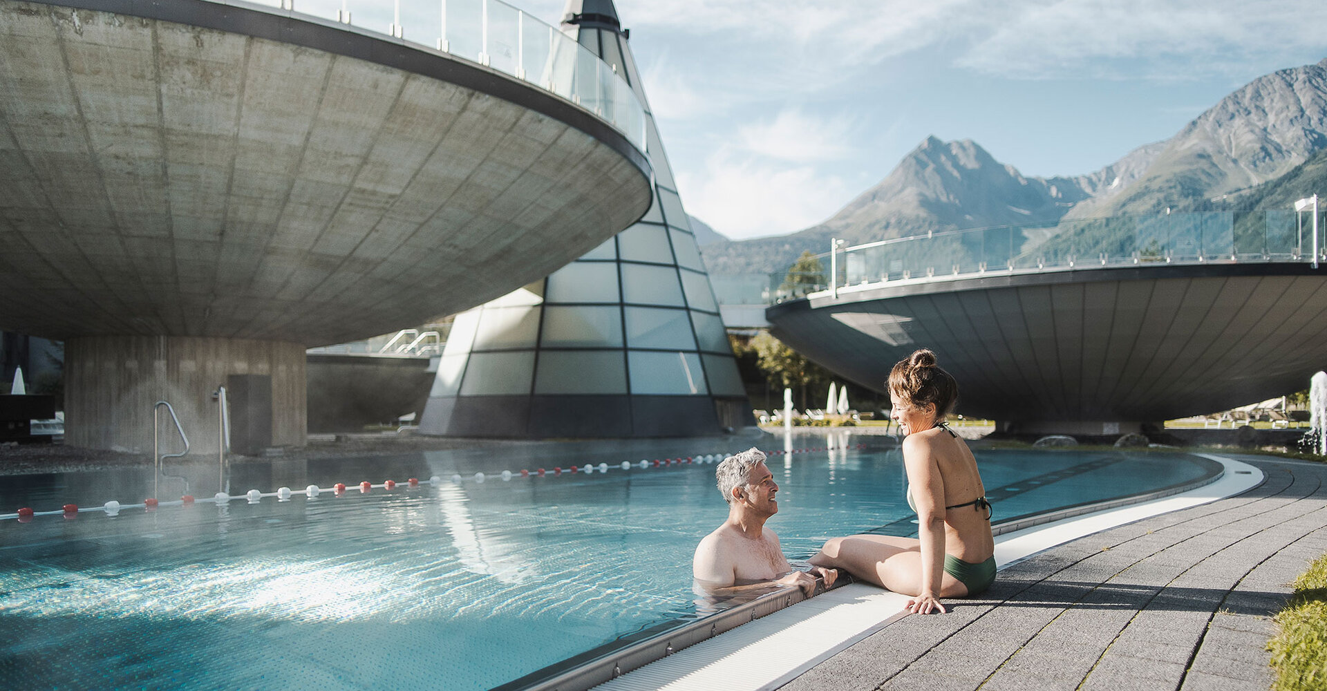 Ein älterer Mann im Schwimmbecken unterhält sich mit einer Frau, die am Beckenrand sitzt. Im Hintergrund sind moderne, runde Betongebäude und Berge vor einem blauen Himmel zu sehen.