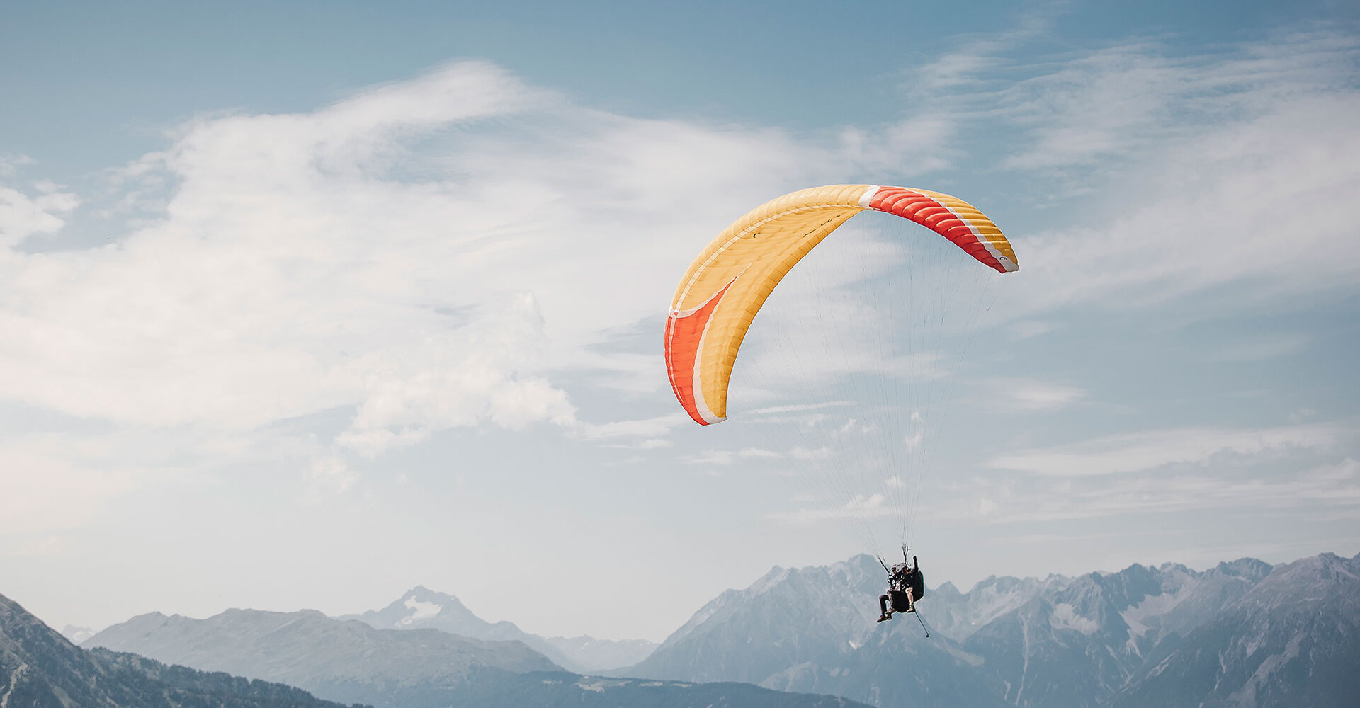 Eine Person fliegt mit einem gelb-orangefarbenen Gleitschirm über den Bergen unter einem teilweise bewölkten Himmel.
