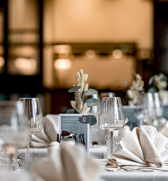 An elegant table setting featuring folded napkins, clear wine glasses and a small floral arrangement, prepared for a festive event or reception in a softly lit dining room.