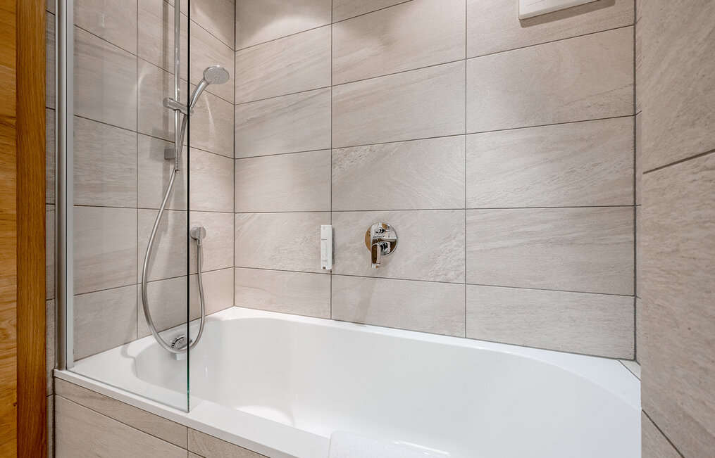 Modernes Badezimmer mit einer weißen Badewanne, einer an der Wand montierten Duschbrause und großen, rechteckigen, beigen Fliesen, die die Wände und die Verkleidung der Badewanne bedecken. Es gibt eine Duschwand aus Glas und verchromte Armaturen.