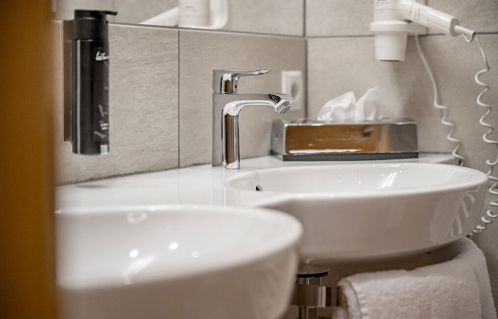 A close-up of a modern washbasin featuring a chrome faucet, a soap dispenser and a wall-mounted hair dryer with white tiled walls and a towel.