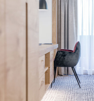 A modern workspace featuring a wooden desk, a black chair with a red seat, a pendant light hanging above and floor-length curtains covering a large window.