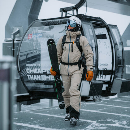 A skier in beige ski gear, wearing a helmet and goggles, carries his skis as he steps out of the mountain gondola cabin on a snowy day.