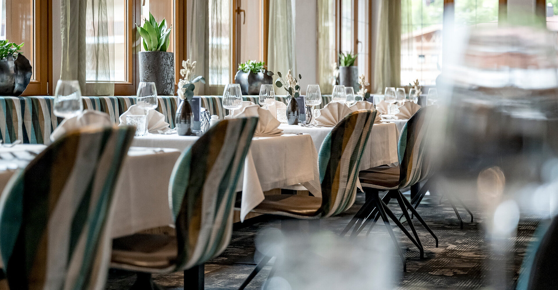 An elegant restaurant with striped chairs, tables covered with white tablecloths, folded napkins, glasses and ornamental plants by large windows through which daylight streams in.