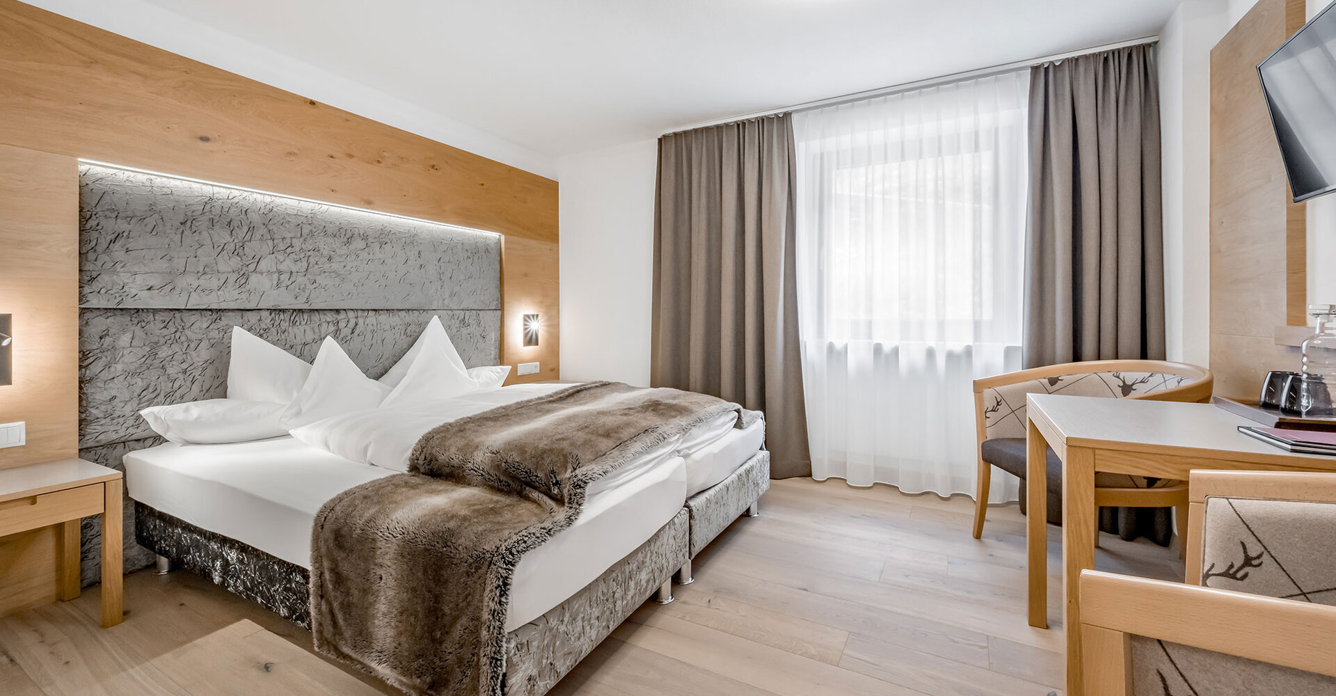 Modern hotel room featuring a double bed, wood accents, a desk with a chair, a wall-mounted TV and a large window with sheer curtains that allow daylight to stream in.