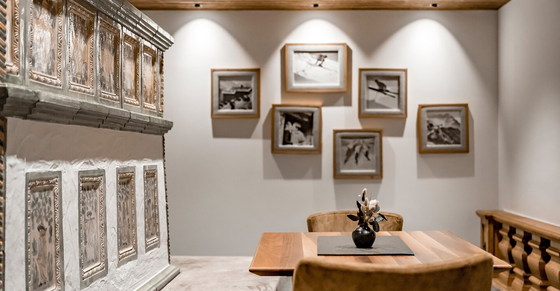 A cozy room with a wooden table, two chairs, a decorative tiled stove and framed black and white photos on a softly lit wall. A small vase of flowers stands on the table.