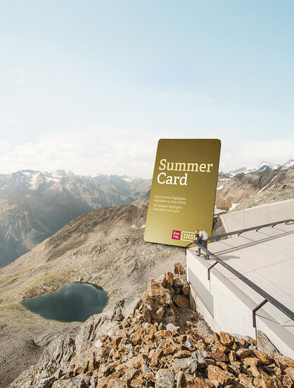 Two people stand on a platform next to a huge "Summer Card" sign, looking out at a small lake and rugged mountain peaks under a clear sky.