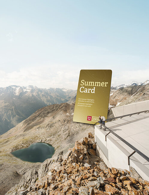 Zwei Personen stehen auf einer Plattform neben einem riesigen "Summer Card"-Schild und blicken unter klarem Himmel auf einen kleinen See und schroffe Berggipfel.