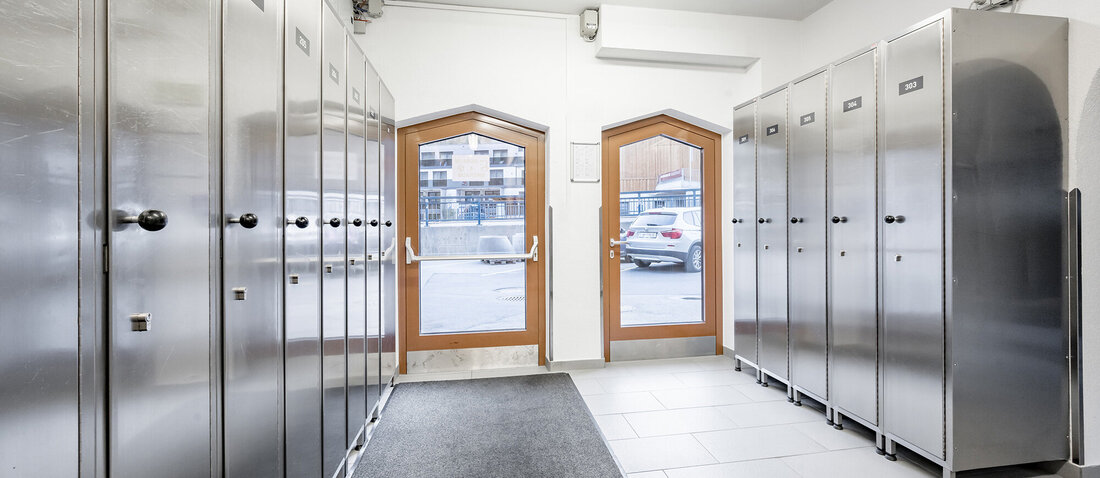 A clean ski storage room with metal lockers on both sides, a black carpet down the middle and two wood-framed glass doors leading outside.
