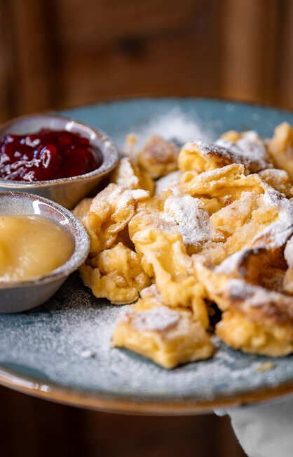 Ein Teller mit Kaiserschmarrn, bestäubt mit Puderzucker, serviert mit kleinen Schälchen mit roter Fruchtkonfitüre und Apfelmus.