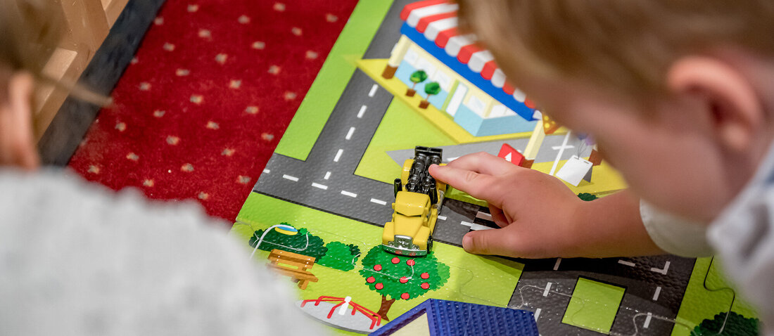 Two children are playing on a colorful play mat featuring streets and buildings, pushing a small yellow toy car along the road.