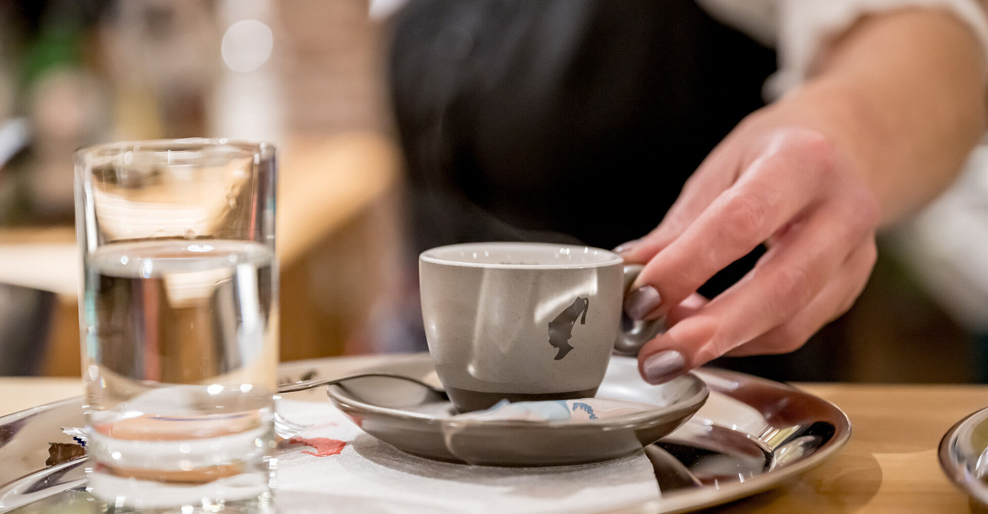A hand places a grey coffee cup on a saucer next to a glass of water on a metal tray, under which lies a napkin at a cafe counter.