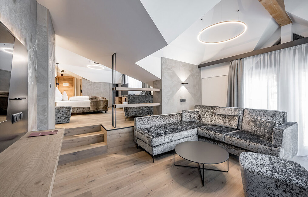 Modern loft-style suite featuring a comfortable grey corner sofa, a round coffee table and an open sleeping area on a raised platform. Large windows with sheer curtains let in plenty of natural light.