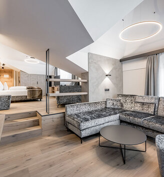 Modern loft-style suite featuring a comfortable grey corner sofa, a round coffee table and an open sleeping area on a raised platform. Large windows with sheer curtains let in plenty of natural light.
