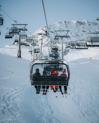 Four people are riding on a ski lift up a snow-covered mountain, surrounded by snowy slopes and other ski lifts under a brilliant blue sky.