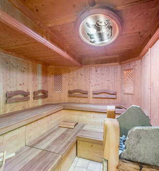 A wood-paneled sauna with tiered wooden benches, a stone heater in the corner and a decorative ceiling light that creates a warm and inviting atmosphere.