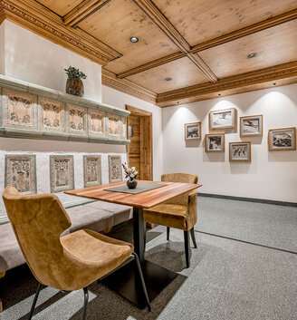 A cozy lounge with wood paneling, a table with two suede chairs, framed pictures on the walls, a mirrored wall reflecting the room and warm mood lighting.