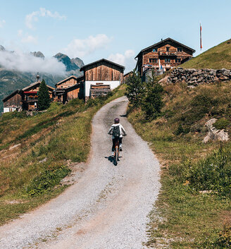 A person is riding a bicycle up a gravel path towards wooden huts on a grassy slope with mountains and a blue sky in the background.