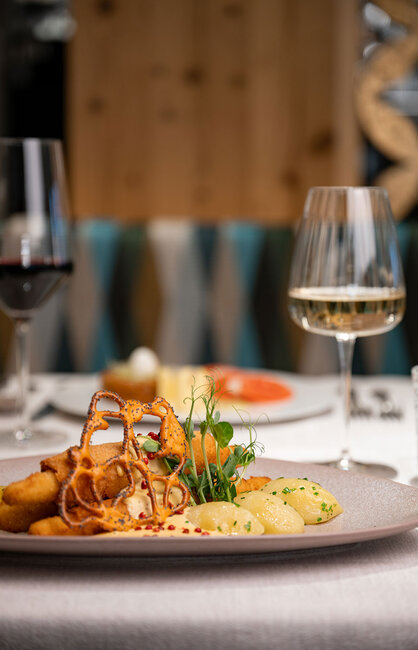 Ein auf einem Teller angerichtetes Gourmetgericht mit gegrilltem Spargel, Kartoffeln und Beilage, serviert auf einem Tisch mit Gläsern Rot- und Weißwein in stilvollem Restaurantambiente.