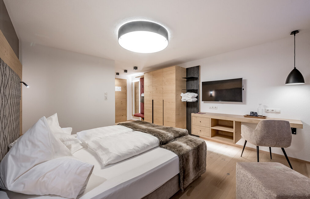 A modern hotel room featuring a large bed, a faux-fur throw, a desk, a chair, a wall-mounted TV, a wardrobe and minimalist lighting. The décor is characterized by neutral tones and clear lines.