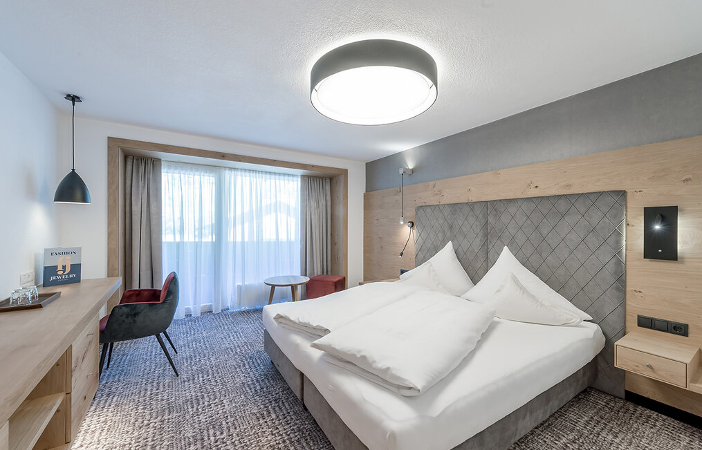 Modernes Hotelzimmer mit einem großen Bett, Holzmöbeln, einem Schreibtisch mit Stuhl, einem kleinen runden Tisch und einem Fenster mit transparenten Vorhängen, durch die Tageslicht hereinfällt.