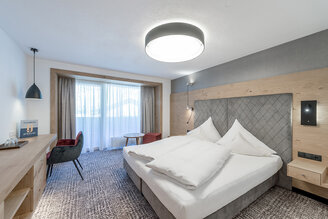 Modern hotel room with a large bed, wooden furniture, a desk with a chair, a small round table and a window with sheer curtains that let in daylight.