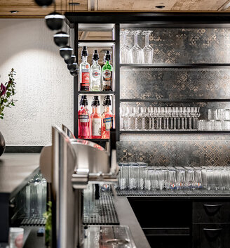 A modern bar featuring glasses and spirit bottles neatly arranged on shelves, a vase of flowers on the counter and a metal beer tap in the foreground. The lighting highlights the clean, stylish design.