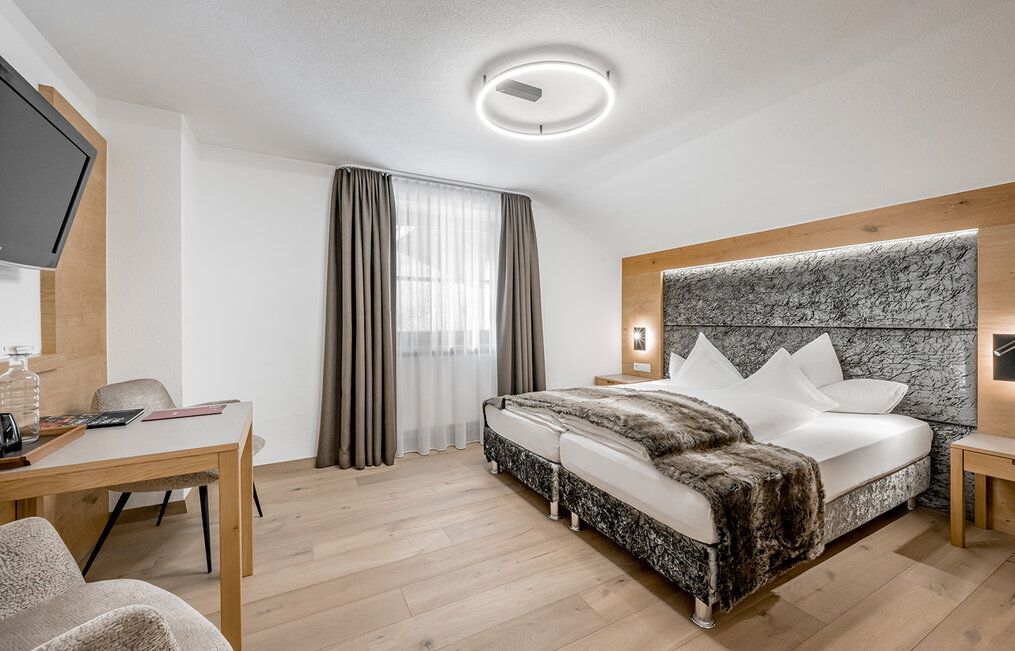 A modern hotel room featuring a large bed, a brown faux-fur throw, wood accents, a wall-mounted TV, a desk, two chairs and a window with white sheer curtains and dark drapes. A round ceiling light illuminates the room.