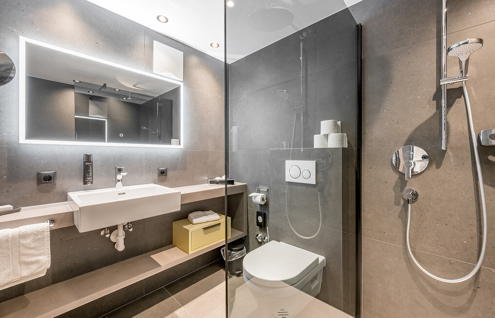 Modern bathroom featuring a wall-mounted washbasin, a toilet, an illuminated mirror, a glass shower enclosure and a rainfall shower, as well as shelving for towels and toilet paper. The walls and floor are clad in gray tiles.