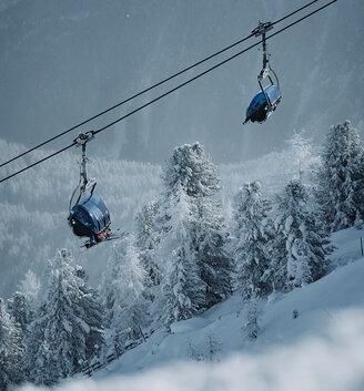 Two blue chairlift seats glide over snow-covered trees on a mountainside with a misty, wooded landscape stretching out in the background