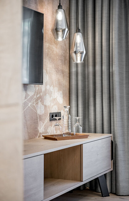 Modernes Sideboard mit zwei darüber hängenden Pendelleuchten, einem Tablett mit zwei Gläsern und einer Glaswasserflasche, grauen Vorhängen und einem Teil eines Fernsehbildschirms an der Wand.
