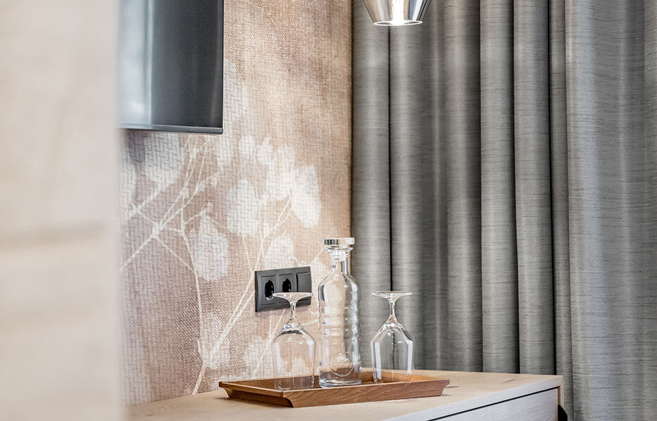 Modern sideboard with two pendant lights hanging above it, a tray with two glasses and a glass water bottle, gray curtains and part of a TV screen on the wall.