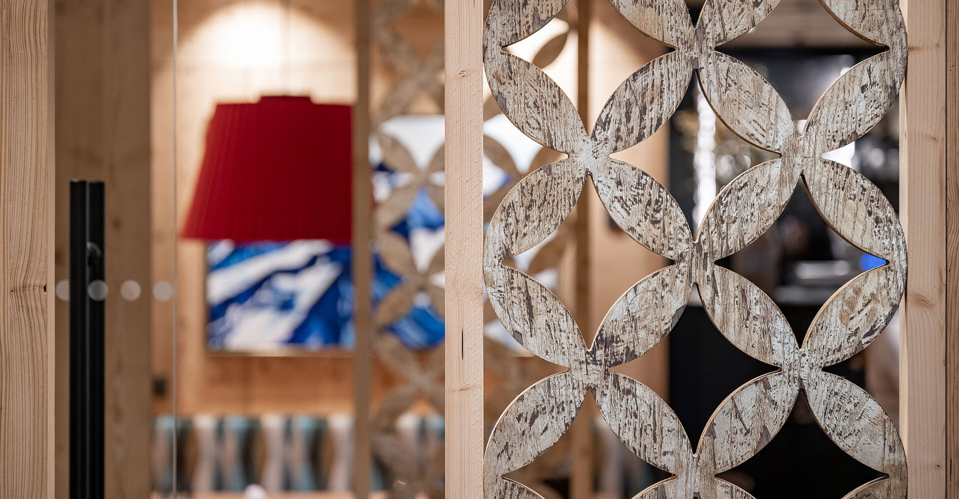 A decorative wooden partition element with a geometric floral pattern stands in the foreground. In the background, a red lampshade, abstract blue-and-white wall art and blurred seating create a cozy living atmosphere.
