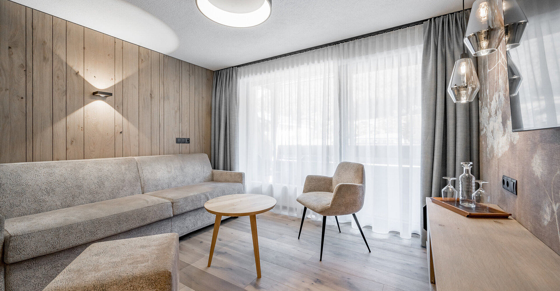 Modern living room with light wood walls and floors, a beige sofa, a matching armchair, a round wooden coffee table and pendant lights. A large window with sheer curtains lets in daylight.