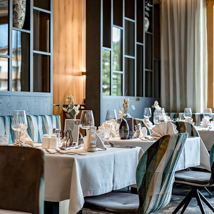 Elegant restaurant interior with neatly set tables, striped upholstered chairs, decorative shelves and large windows with light curtains through which daylight streams in.