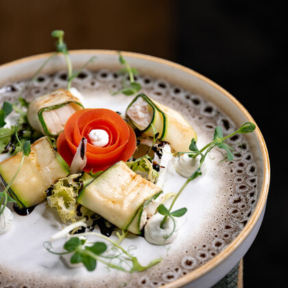 A gourmet meal consists of cheese-wrapped packets, a tomato rose centerpiece, green garnish and small dollops of dip, beautifully arranged on a textured ceramic plate.