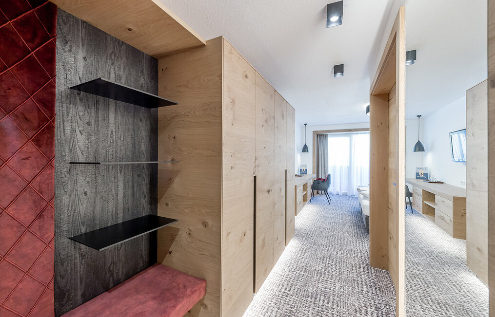 Modernes Hotelzimmer mit hellen Holzvertäfelungen, offenen Regalen, einer roten gepolsterten Bank, Teppichboden, einem Schreibtisch mit Stuhl und einem großen Fenster mit transparenten Vorhängen, durch die Tageslicht hereinströmt.