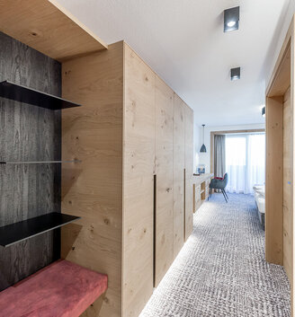 Modern hotel room with light wood paneling, open shelving, a red upholstered bench, carpeting, a desk with a chair and a large window with sheer curtains allowing daylight to stream in.