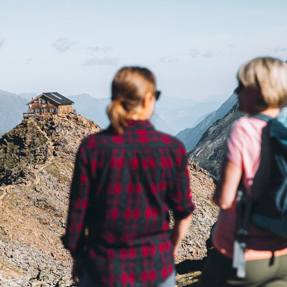 Two people carrying backpacks stand on a rocky mountain path and look at a wooden hut perched on a distant mountain ridge, surrounded by mountain peaks under a clear sky.