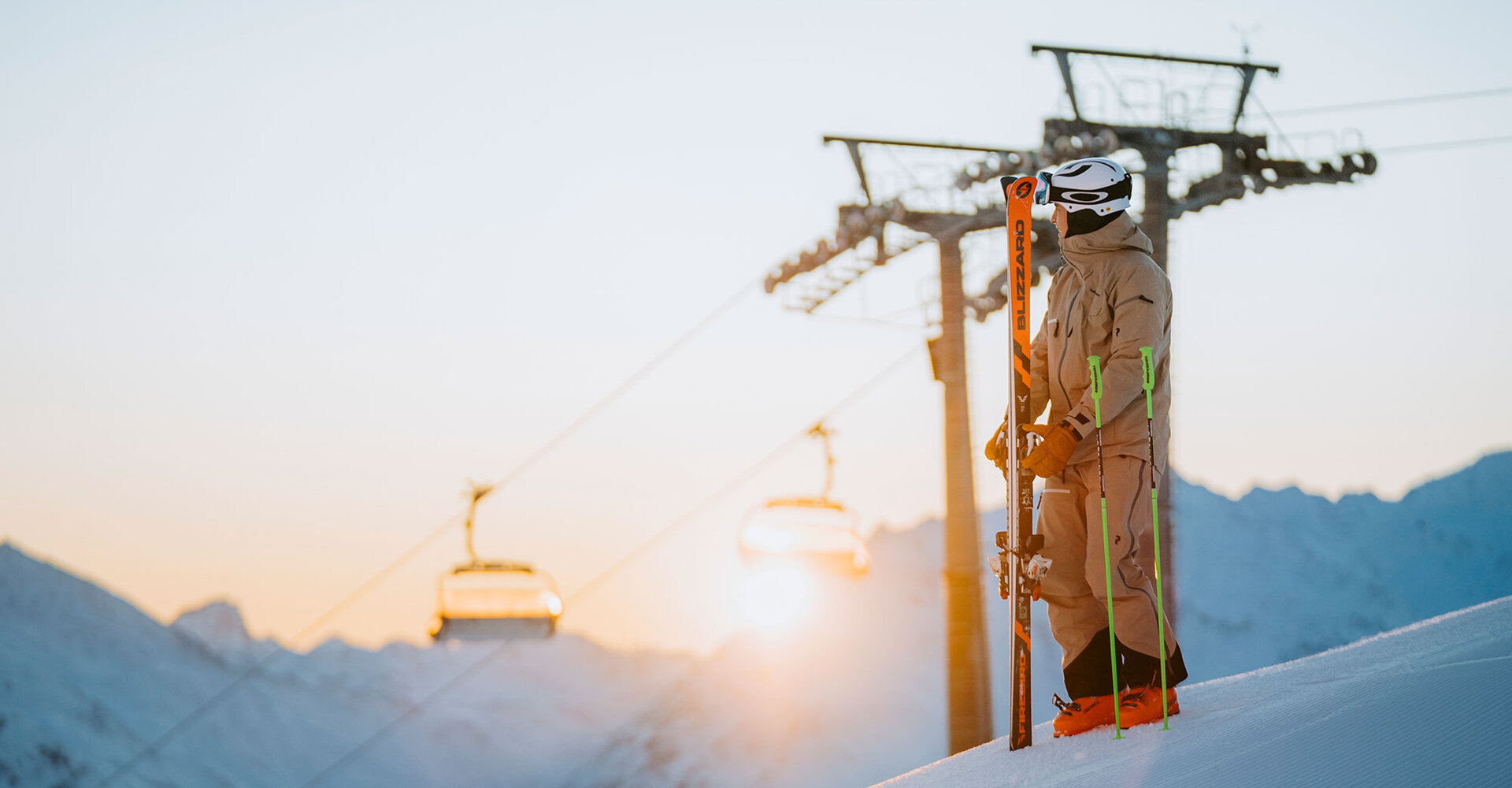 A skier in beige clothing stands on a snowy slope at sunset, holding his skis. In the background, ski lifts and mountain peaks are under a clear sky.