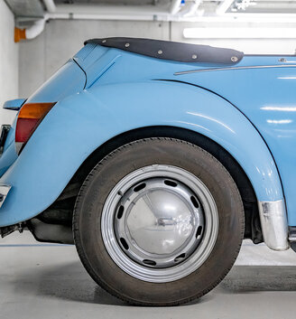 A close-up, side-view shot of the rear half of a light blue vintage convertible parked in a garage: the wheel, tire, bumper and fabric soft top are in the picture.