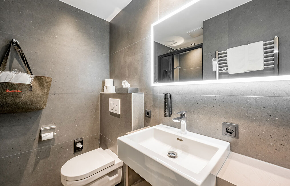 Modern bathroom with grey-tiled walls, a wall-mounted toilet, a white washbasin with a chrome faucet, a large illuminated mirror, a towel holder with a white towel and a hanging felt bag. The shower area is in the background.