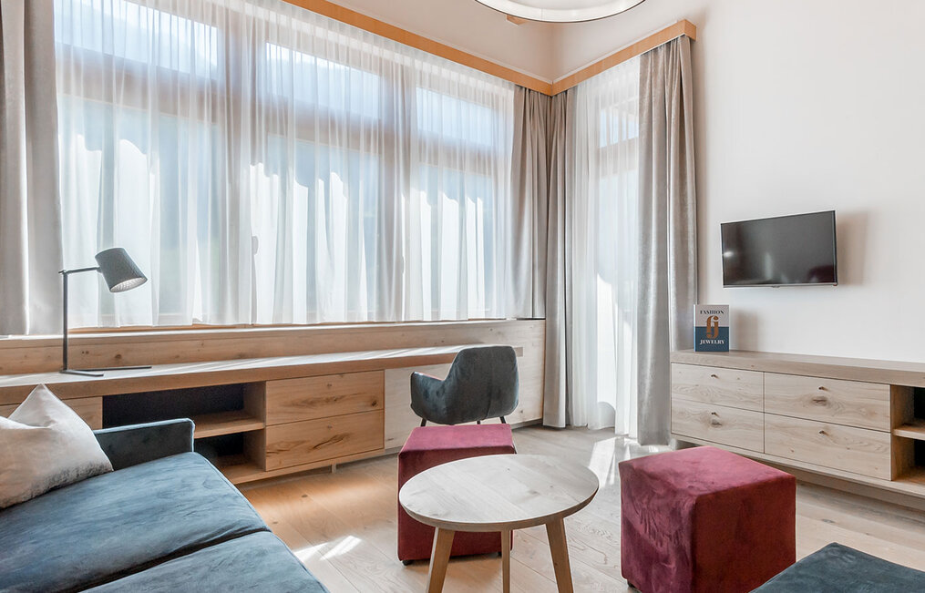 Bright, modern living room with large windows, sheer curtains, a gray sofa, a wooden desk and cabinets, a round coffee table, an armchair, two red stools, a floor lamp and a wall-mounted TV.