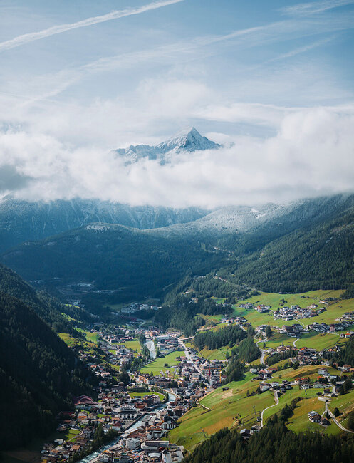 Aerial view of a village nestled in a green valley, surrounded by wooded hills and high mountains with a layer of clouds and snow-capped peaks in the background under a blue sky.