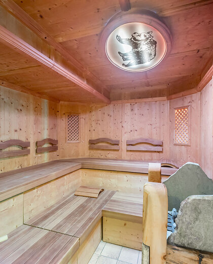 Ein mit Holz vertäfelter Saunaraum mit gestuften Holzbänken, einem Steinofen in der Ecke und einer runden Deckenleuchte mit einem dekorativen Motiv.