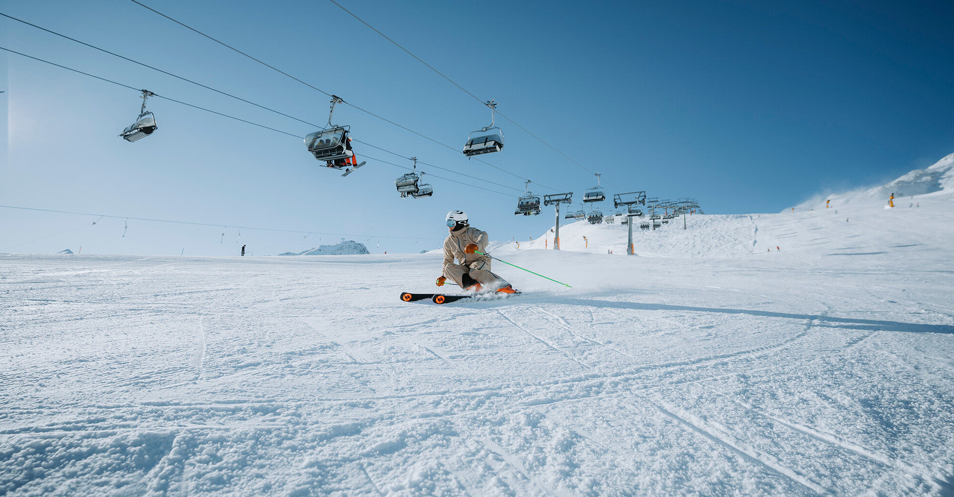 A skier in winter clothing speeds down a snowy slope under a bright blue sky, while in the background several ski lifts transport people.