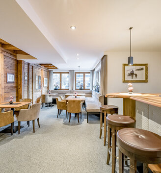 Modern lounge with wood accents, light brown chairs, small tables, a bar with bar stools and large windows allowing daylight to stream in. Cozy, inviting atmosphere.