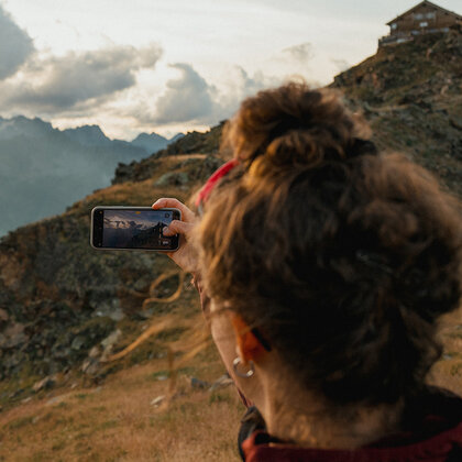 A person with curly hair is photographing a mountain landscape at sunset using a smartphone. The scene shows grassy hills, rocky terrain and a hut on the hillside under a partly cloudy sky.