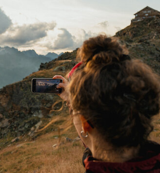 A person with curly hair is photographing a mountain landscape at sunset using a smartphone. The scene shows grassy hills, rocky terrain and a hut on the hillside under a partly cloudy sky.