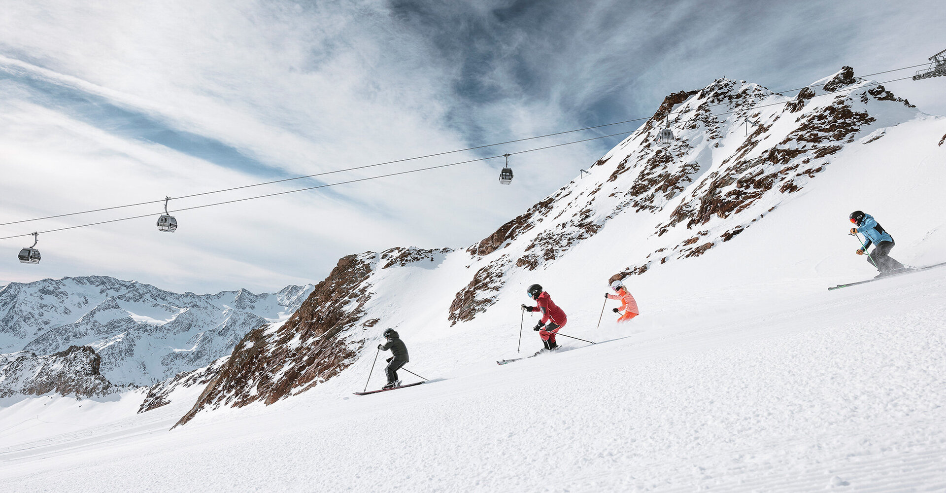 Four people are skiing down a snow-covered mountain slope under a partly cloudy sky while a ski lift and snow-capped peaks are in the background.